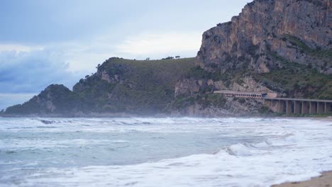 Picturesque-beach-view-of-white-ocean-waves-rolling-towards-and-breaking-on-sandy-beach-with-bridge-tunnel-and-mountain-in-background,-Gaeta,-Italy,-static