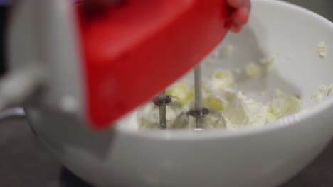 close-up-of-woman-using-a-red-hand-mixer-to-mix-cheesecake-ingredients-in-a-white-bowl-in-a-kitchen