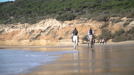 Handheld-low-angle-shot-on-beach-with-calm-waves-in-blue-sea-with-view-of-two-riders-with-their-horses-riding-along-the-beach