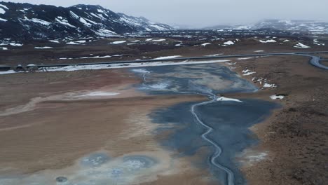 Aerial-panoramic-shot-of-geothermal-mud-pots-with-snowy-mountains-in-background-in-Iceland