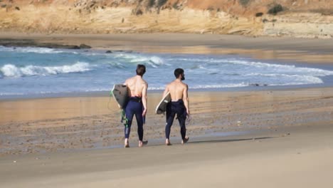 Handheld-shot-of-two-male-sporty-surfers-with-their-surfboards-running-along-a-charming-sandy-beach-while-calm-waves-in-blue-sea