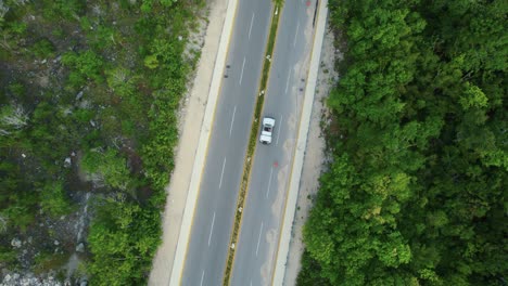 Aerial-drone-shot-of-a-person-dancing-on-a-white-cabriolet-car,-driving-on-a-empty-asphalt-road,-going-through-palm-trees,-in-Tulum,-Mexico
