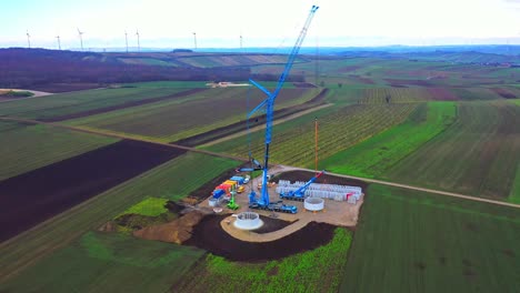 Crane-At-The-Construction-Site-Of-Wind-Turbine-Surrounded-By-Fields