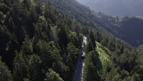 Reveal-of-red-minivan-driving-along-a-steep-mountain-through-forest-in-the-Alpes-on-a-sunny-morning---drone-follow-cam