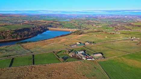 Aerial-drone-footage-of-traffic-on-a-country-road-with-the-M62-motorway-in-the-distance-on-Scammonden-water-with-farmland-lake,-reservoir,-and-wide-open-views-of-the-Pennine-Hills