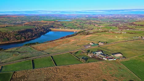 Aerial-drone-footage-of-traffic-on-a-country-road-with-the-M62-motorway-in-the-distance,crossing-Scammonden-water-with-farmland-lake,-reservoir,-and-wide-open-views-of-the-Pennine-Hills