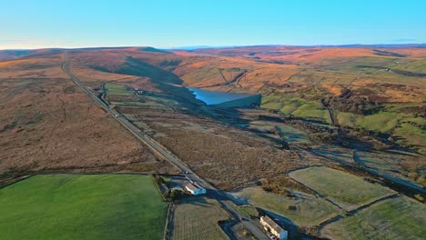 Aerial-drone-footage-of-traffic-on-a-country-road-on-Saddleworth-Moor-with-golden-land-lake,-reservoir,-and-wide-open-views-of-the-pennines