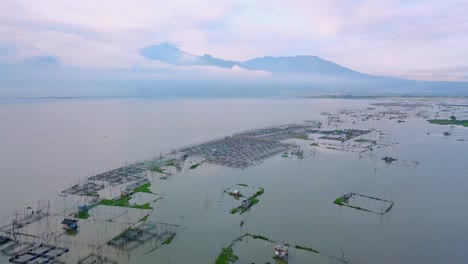 Aerial-forward-flight-over-fishing-farm-with-cages-in-RAWA-PENING-LAKE,-INDONESIA---Clouds-covering-mountains-in-background