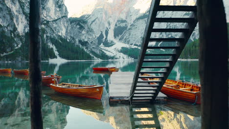 Passing-under-a-boathouse-and-out-over-a-dock-and-across-the-pristine-waters-of-Lago-di-Braies,-Italy,-aerial