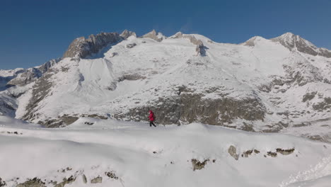 Toma-Aérea-En-Suiza-Con-Una-Persona-Caminando-Con-Raquetas-De-Nieve-En-Un-Día-Soleado-Con-Un-Glaciar-Detrás