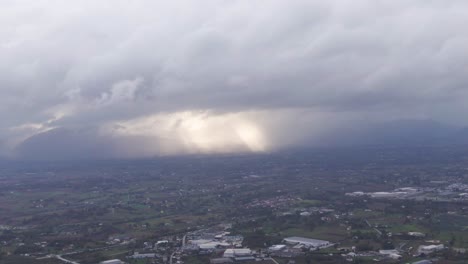 Ominous-grey-stormy-clouds-blanket-flat-countryside-landscape-below-with-sun-rays-beaming-in-background-through-hole-in-sky,-aerial