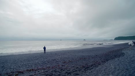 Person-watches-ocean-on-grey,-moody-beach,-runs-from-waves-on-shore