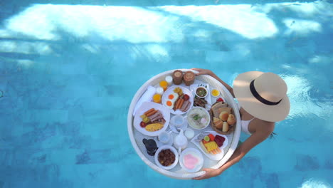 Overhead-view-of-a-female-tourist-walking-in-a-crystal-clear-pool-with-a-floating-breakfast-with-delicious-food-on-it