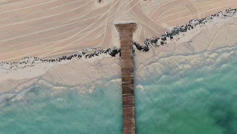 AERIAL-DIRECTLY-ABOVE-Wooden-Jetty-With-Clear-Water-Lapping-At-Sandy-Beach