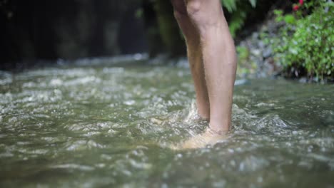 A-male-European-hiking-tourist-is-standing-with-bare-feet-in-cold-water-creek-a-river-close-to-a-large-waterfall-cliff-in-the-green-forest-jungle-of-Tanzania