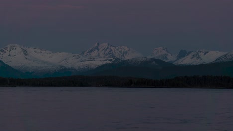 Beautiful-Time-Lapse-of-snow-covered-mountains-in-the-background-and-ocean-in-the-foreground,-separated-by-a-forest-during-sunset