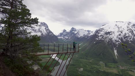Girl-walking-out-to-viewpoint-on-a-metal-bridge,-watching-over-beautiful-scenic-view-of-snowy-mountains,-meadows-and-rivers-in-a-valley