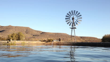 Landwirtschaftliche-Wasserpumpe-Dreht-Sich-In-Einer-Landwirtschaftlichen-Umgebung-Und-Pumpt-Wasser-In-Einen-Lagertank-Gegen-Eine-Trockene-Landschaft-In-Südafrika
