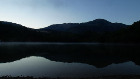 Dark-still-lake-reflective-waters-by-surrounding-mountains-in-countryside-at-sunrise,-static
