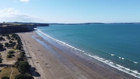 Aerial-shot,-flying-over-a-beautiful-beach-with-people-on-a-sunny-day-in-Long-Bay,-New-Zealand