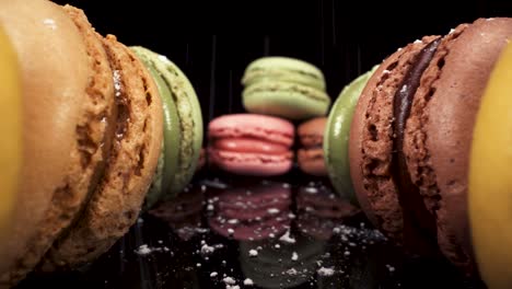 Reverse-moving-macro-view-past-macarons-with-powdered-sugar-falling-on-black-reflective-glass,-luxury-french-pastries,-sweet-sugary-food