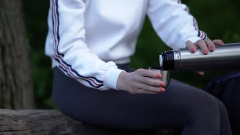 Panning-view-of-young-caucasian-girl-sitting-on-huge-log-in-forest-and-pouring-herself-hot-drink-from-thermos-bottle