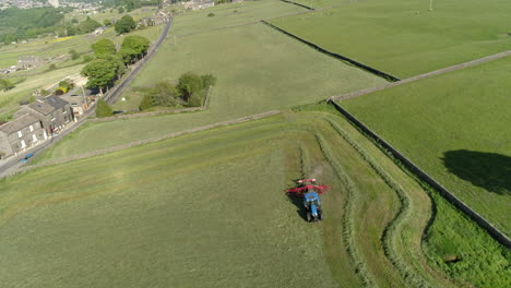 Farmer-Driving-Blue-Tractor-cuts-the-luscious-green-grass-in-a-countryside-farm-meadow-in-West-Yorkshire,-Drone-Aerial-flyover