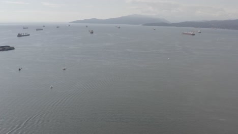 Aerial-panaramic-fly-over-sunny-smog-English-Bay-to-Kitsilano-beach-harbor-where-freightor-barge-vessels-await-industry-export-import---leisure-boats-sail-in-Summer-at-Stanley-Park-party-place-ora2-3