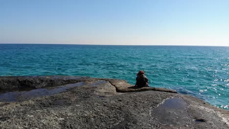 Aerial-tracking-shot-of-girl-sitting-on-rock-looking-at-blue-water-with-wind-blowing-her-hair