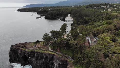The-Wonderful-View-Of-Kadowaki-Lighthouse-In-Japan-With-A-Beautiful-Coastline-And-Green-Trees-Under-The-Cloudy-Sky---Aerial-Tilting-up-Shot