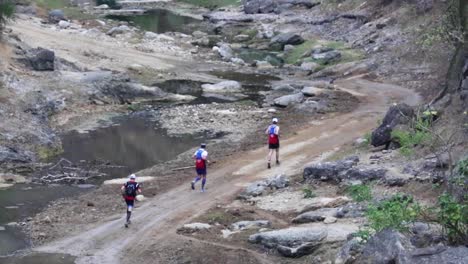 Gruppe-Von-Marathonathleten,-Die-Ein-Wohltätigkeitsrennen-Auf-Einer-Unbefestigten-Straße-Entlang-Eines-Flusses-Durch-Einen-Walddschungel-In-Der-Ländlichen-Wunderschönen-Natur-Ecuadors-Laufen-50-Fps