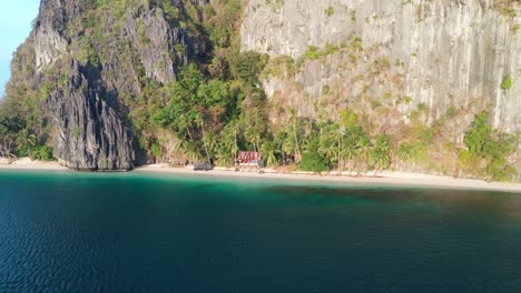 Isolated-Tropical-Beach-With-Cottage-Under-Steep-Cliff,-Pull-Back-Aerial-View-of-Pinagbuyutan-Island,-El-Nido,-Palawan,-Philippines