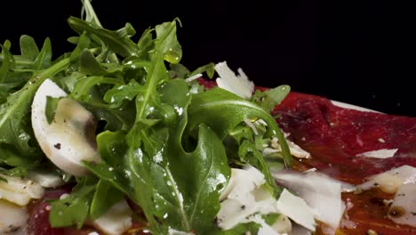closeup-moving-over-macro-view-beef-carpaccio-with-rocket-salad-and-slices-of-Parmigiano-falling-from-the-sky-4k-filmed-on-black-background,-Italian-food-dish