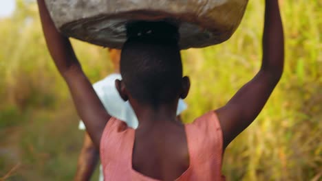Group-of-local-young-African-women-and-kids-are-carrying-big-heavy-buckets-of-drinking-water-and-bringing-the-water-on-their-heads-towards-the-village-walking-in-bush-field---close-shot