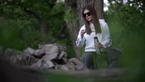Young-caucasian-girl-sitting-in-forest,-pouring-herself-hot-drink-from-thermos-bottle-and-drinking-it