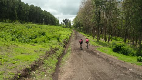 Aerial-drone-shot-flying-above-marathon-athletes-on-mountainbikes-cycling-on-a-dirt-road-through-grass-field-and-farmland-very-heavy-workout-in-jungle-forest-Tanzania-following-shot-4k