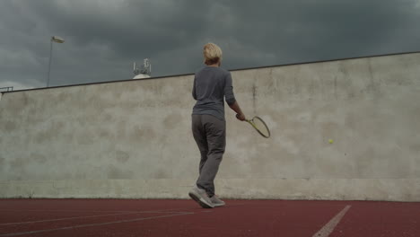 Woman-hits-tennis-ball-against-wall-under-storm-clouds,-low-angle