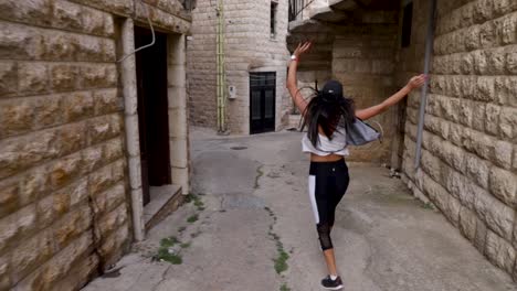 Young-Caucasian-Lebanese-woman-jumping-and-skipping-happily-in-narrow-stone-wall-city-pathway,-behind-close-up-pan