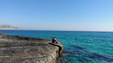 Aerial-tracking-shot-of-girl-sitting-on-a-rock-reading-a-book-with-beautiful-blue-water-behind