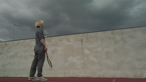 Woman-practices-tennis-against-wall-as-storm-clouds-gather,-low-angle