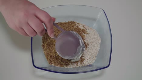 Adding-flour-to-flax-and-quick-oats-in-glass-mixing-bowl,-Overhead-Closeup