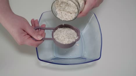 Measuring-rolled-oats-from-glass-jar-into-mixing-bowl,-Overhead-Closeup