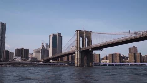 Brooklyn-Bridge-from-East-River-with-the-view-of-Manhattan,-Financial-District-in-slow-motion-during-sunny-weather-at-noon