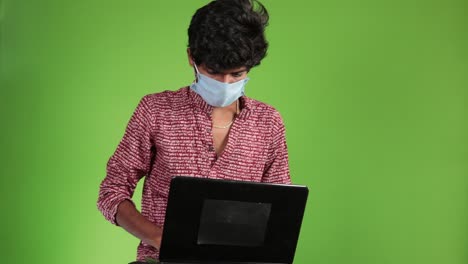 Young-happy-businessman-in-a-red-shirt-with-mask-works-on-a-laptop-on-a-green-screen