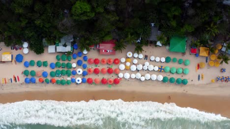 Gorgeous-aerial-drone-bird's-eye-top-view-flyby-of-the-tropical-beach-Praia-do-Madeiro-with-colorful-umbrellas,-clear-blue-water,-tourists-swimming,-and-surfers-near-Pipa-in-Northeastern-Brazil