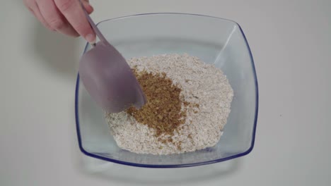 Pouring-half-cup-of-milled-flaxseed-on-top-of-rolled-oats-in-glass-bowl,-Closeup
