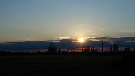 Hazy-Sun-Setting-Down-Over-Wheat-Field-With-Oil-Refinery-Silhouette-on-Horizon,-Time-Lapse-4K