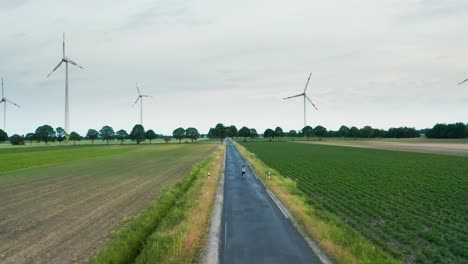 Young-female-is-riding-her-bicycle-on-a-long-road-through-a-green-field-of-windmills-renewable-energy-technology-park-with-tall-turbines-in-dutch-farmland-4k-following-drone-shot