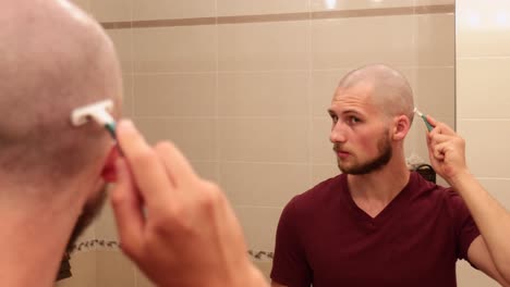 Young-bald-man-shaving-his-own-head-while-looking-in-a-mirror,-view-of-back-but-focus-on-reflection
