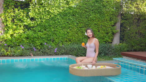 Slow-motion-on-slim-Asian-woman-holding-a-glass-of-orange-juice-and-ready-to-eat-breakfast-from-floating-on-the-water-tray-poolside